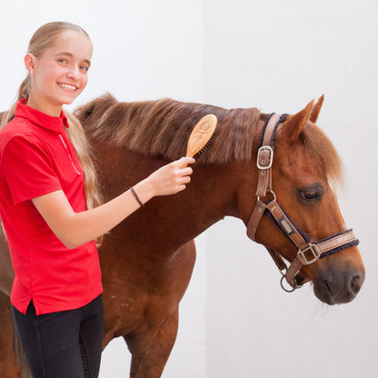 A young girl with blonde hair in a ponytail, wearing a red polo shirt and black pants, smiling as she brushes a brown horse with a wooden paddle brush. The horse is wearing a brown halter.